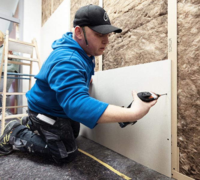 Ein Handwerker montiert eine Trockenbauplatte an einer gedämmten Wandkonstruktion. Sichtbar sind Dämmung, Trockenbauplatte und SORIWA Zellfaserstoffprofile, die Multi Profile.